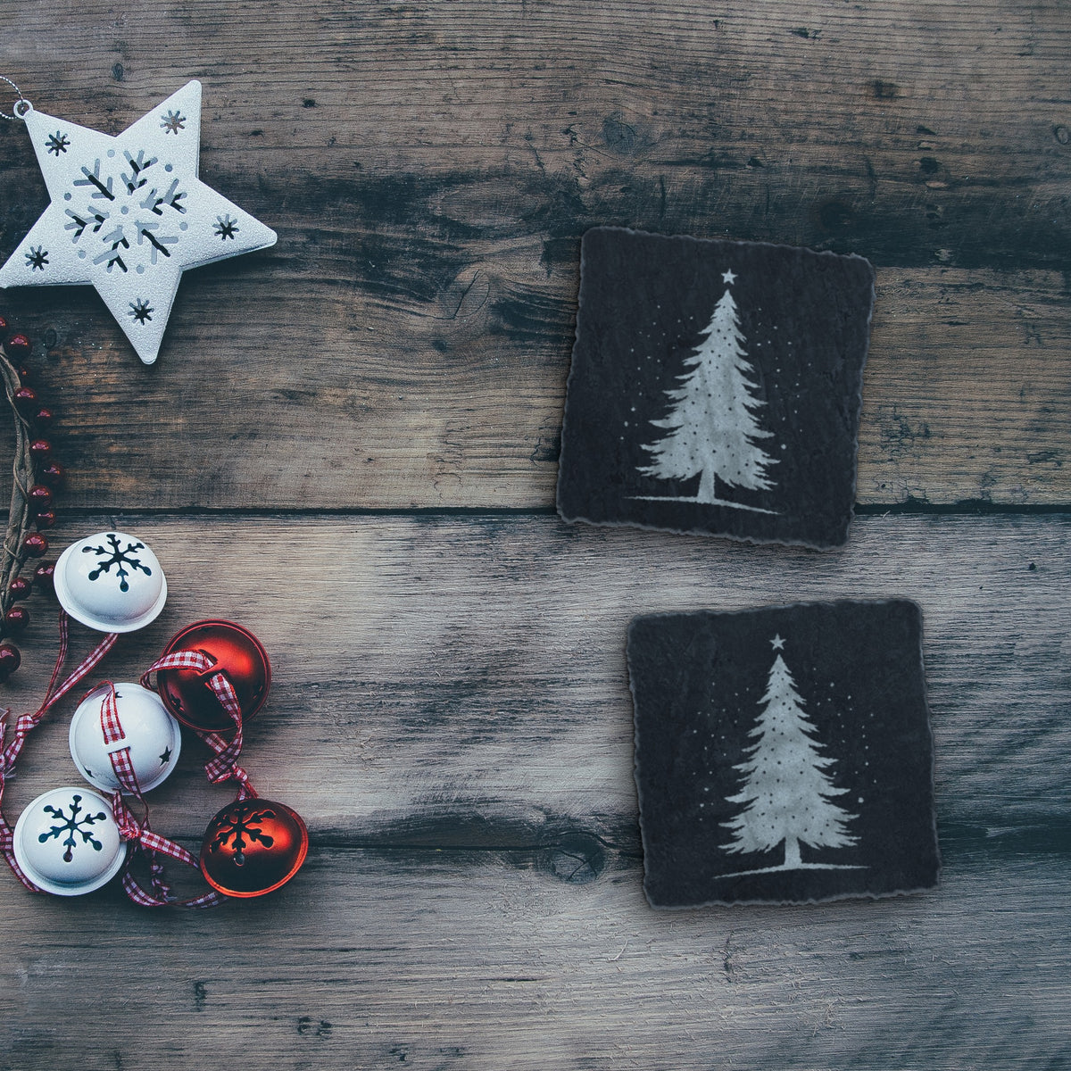 Two snowy Christmas tree slate coasters on a rustic wood table with holiday decorations (product mockup)