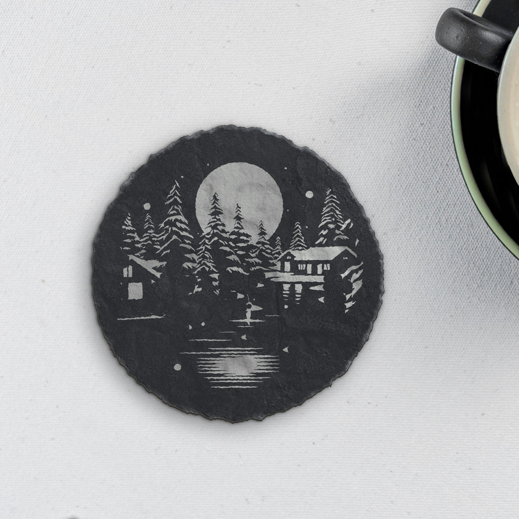 Round slate coaster engraved with a moonlit lakeside cabin among snowy pines (product mockup)