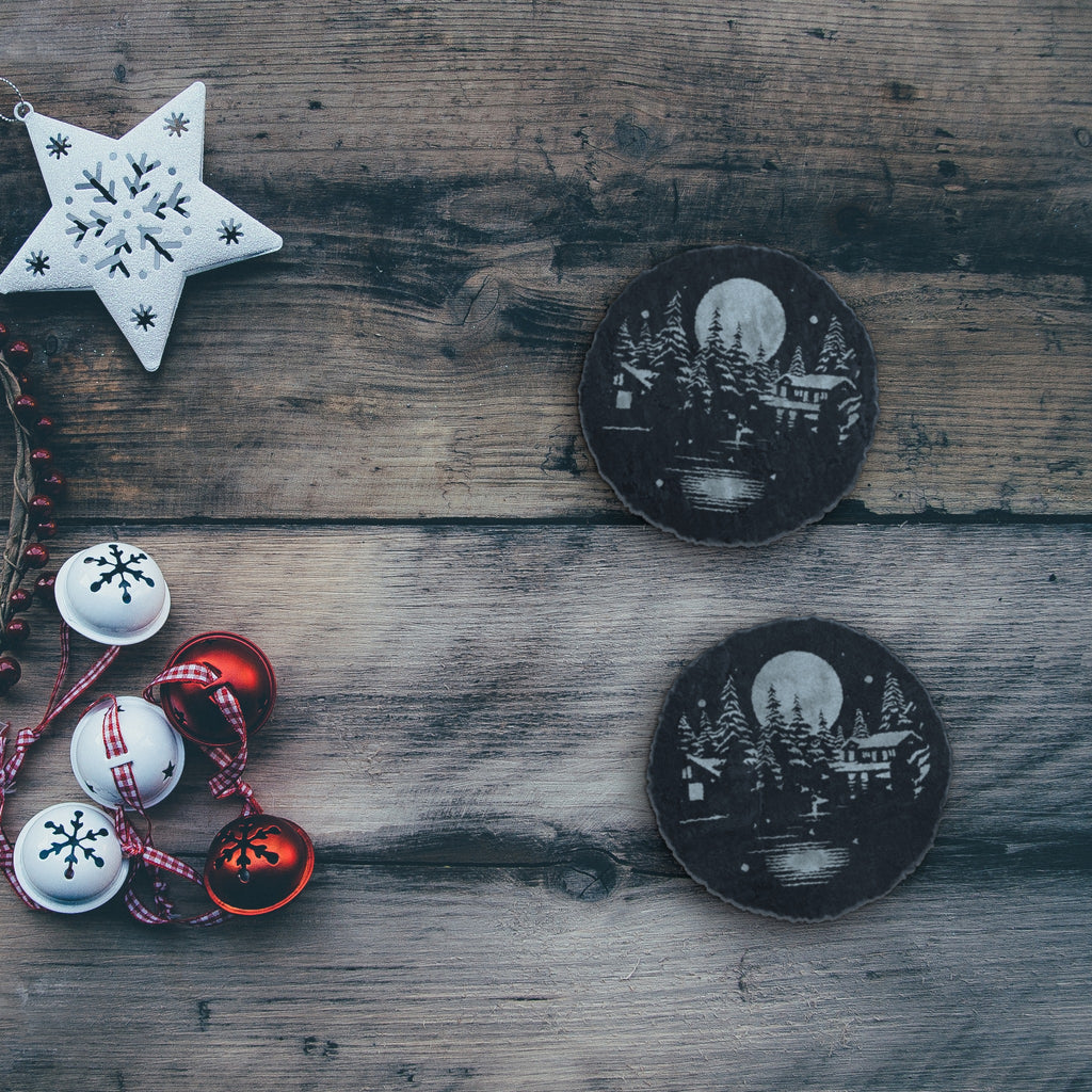 Two winter cabin slate coasters on a rustic wood table with holiday decorations (product mockup)