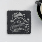 Square slate coaster engraved with a raccoon in a Santa hat holding a sign that says Sorry Santa I've Been Feral (product mockup)