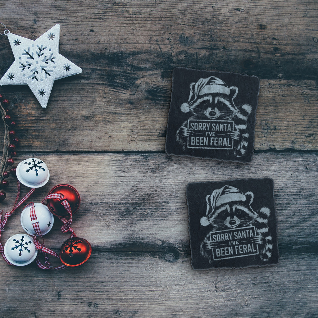 Two feral raccoon Christmas slate coasters on a rustic wood table with holiday decorations (product mockup)