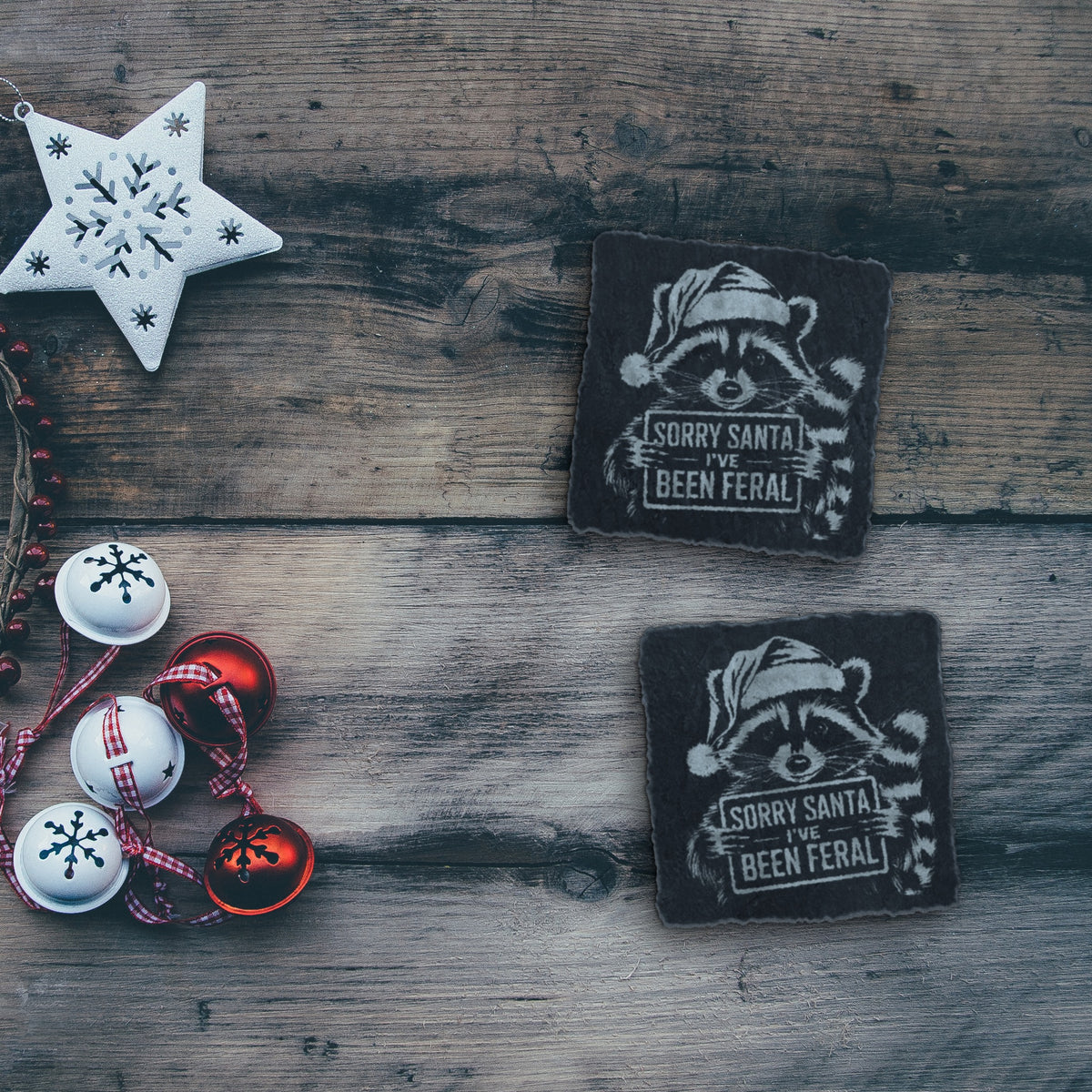 Two feral raccoon Christmas slate coasters on a rustic wood table with holiday decorations (product mockup)