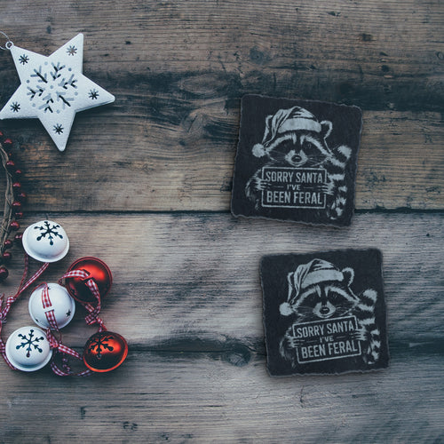 Two feral raccoon Christmas slate coasters on a rustic wood table with holiday decorations (product mockup)