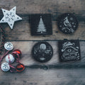 Four Christmas slate coasters, a snowy tree, a winter cabin, a cardinal and a Sorry Santa raccoon, on a rustic wood table with decorations (product mockup)