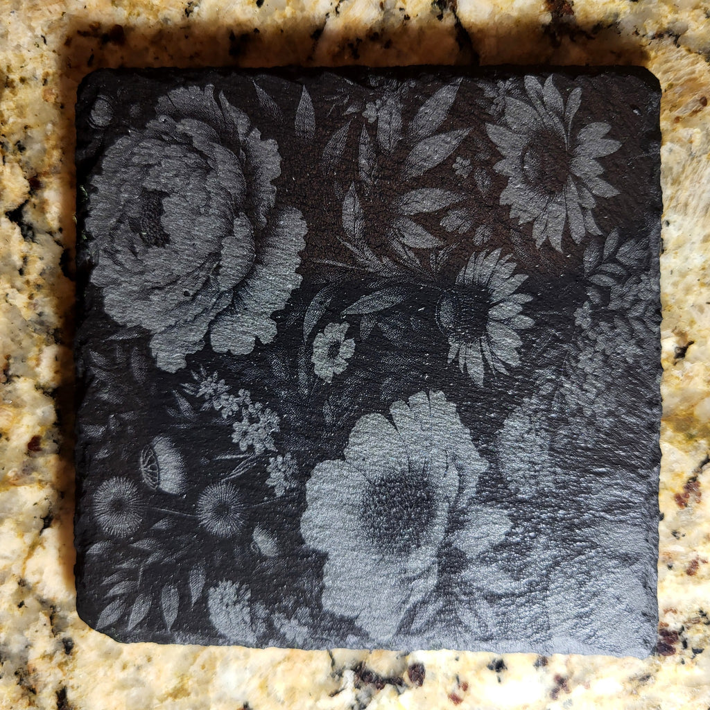 Close-up of a floral engraved slate coaster with peonies and daisies on a granite countertop
