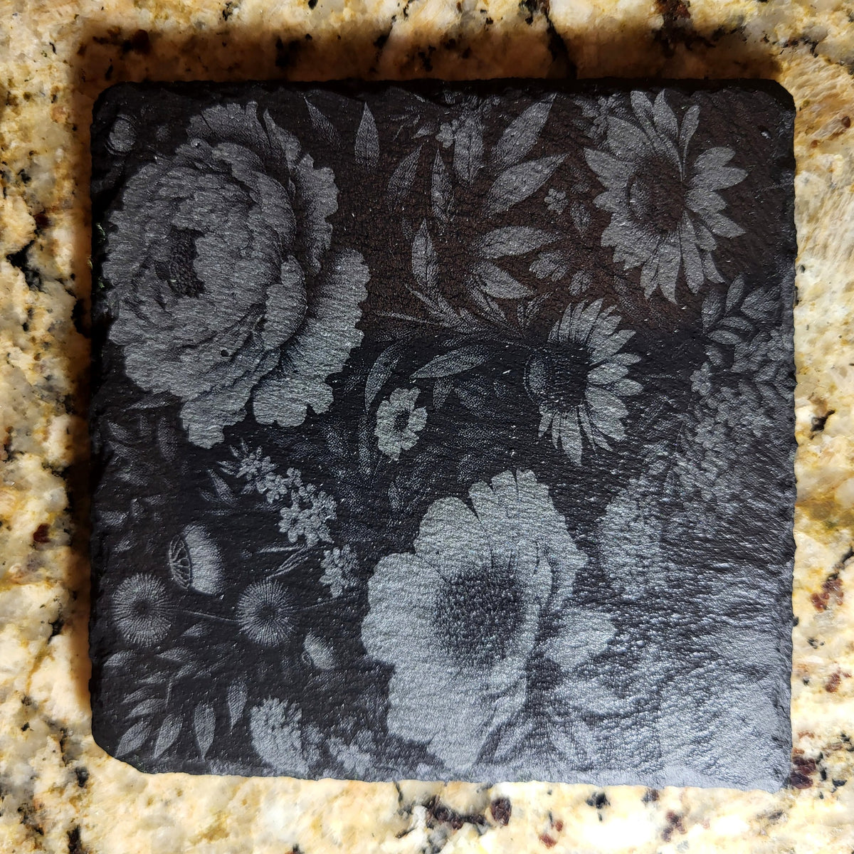 Close-up of a floral engraved slate coaster with peonies and daisies on a granite countertop